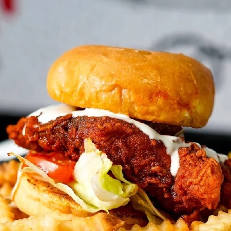 Spicy fried chicken sandwich with lettuce, tomato, and sauce on a bun, served with waffle fries.