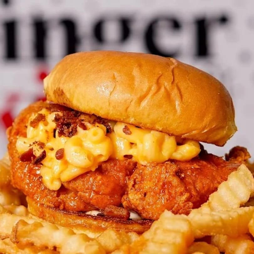 A fried chicken sandwich topped with mac and cheese and bacon bits, served with waffle fries.